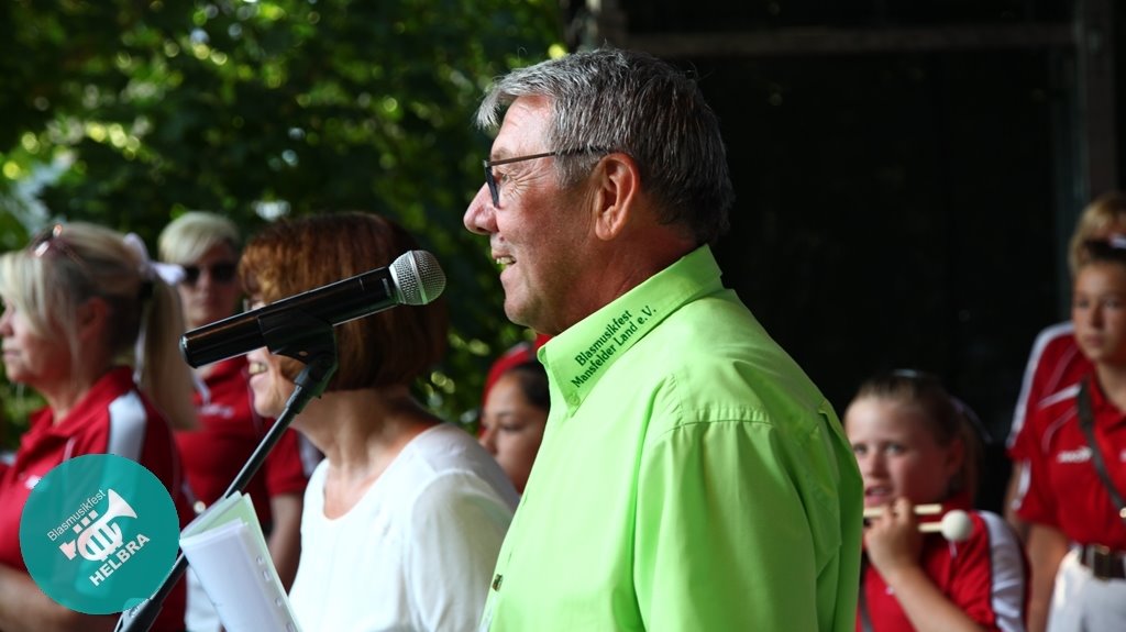Bernd Skrypek im grünen Hemd mit Aufdruck ‚Blasmusikfest Mansfelder Land e.V.‘ spricht in ein Mikrofon.