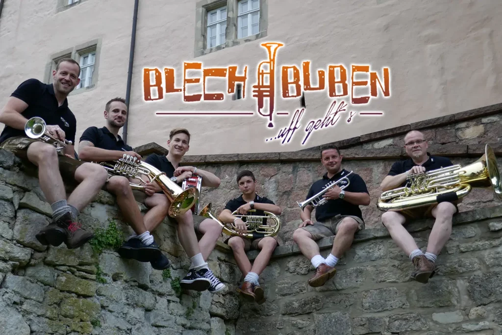 Sechs Musiker der Blechbuben mit Blasinstrumenten auf einer Steinmauer, daneben ihr Logo mit Trompete.