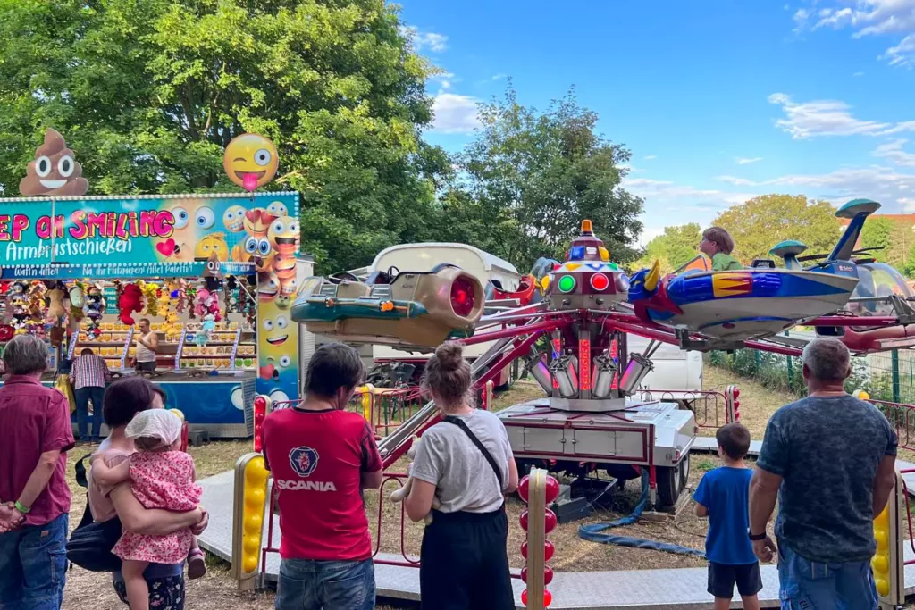 Kinderkarussell auf dem Blasmusikfest, davor Familien mit Kindern, im Hintergrund ein bunter Schießstand mit Smileys.