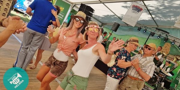 Zwei fröhliche Frauen mit Sonnenbrillen tanzen auf dem Fest, weitere Besucher im Hintergrund.