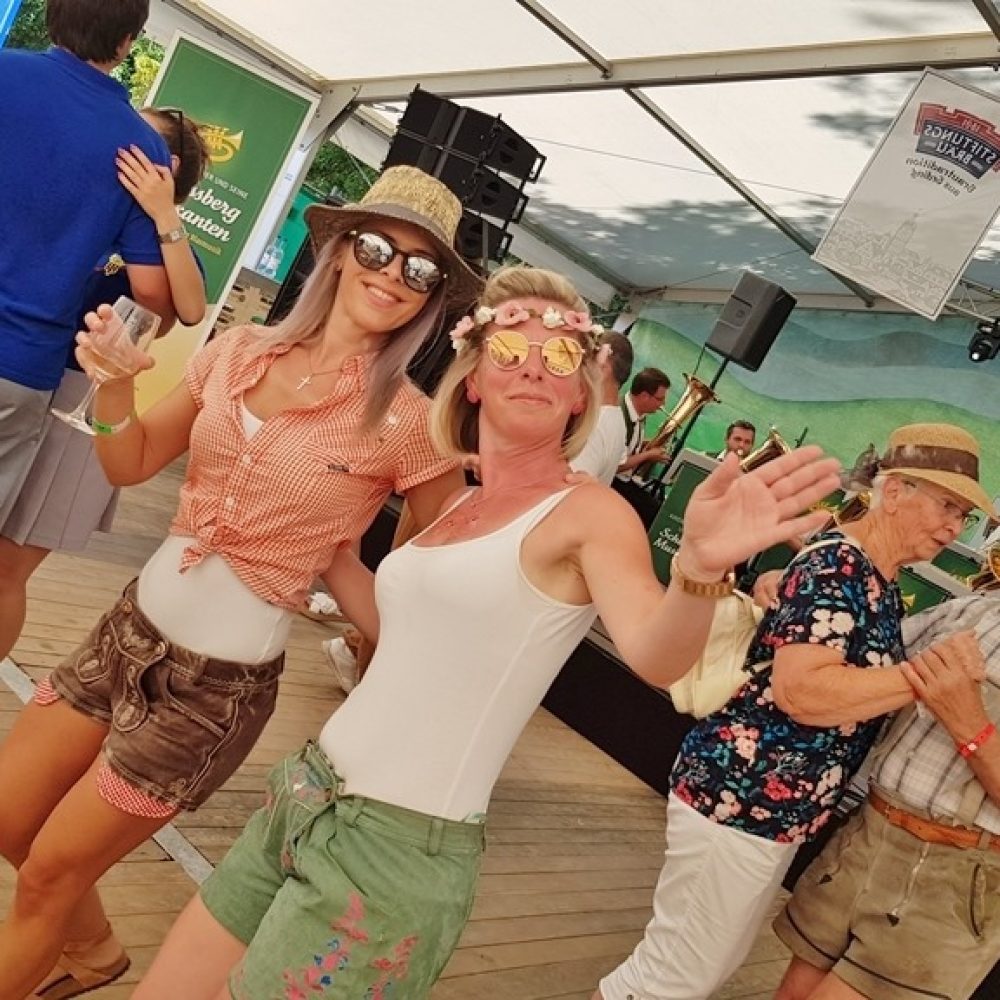 Zwei fröhliche Frauen mit Sonnenbrillen tanzen auf dem Fest, weitere Besucher im Hintergrund.