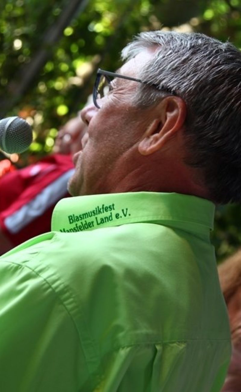 Redner beim Blasmusikfest Mansfelder Land e.V. Bernd Skrypek im grünen Hemd mit Aufdruck ‚Blasmusikfest Mansfelder Land e.V.‘ spricht in ein Mikrofon.